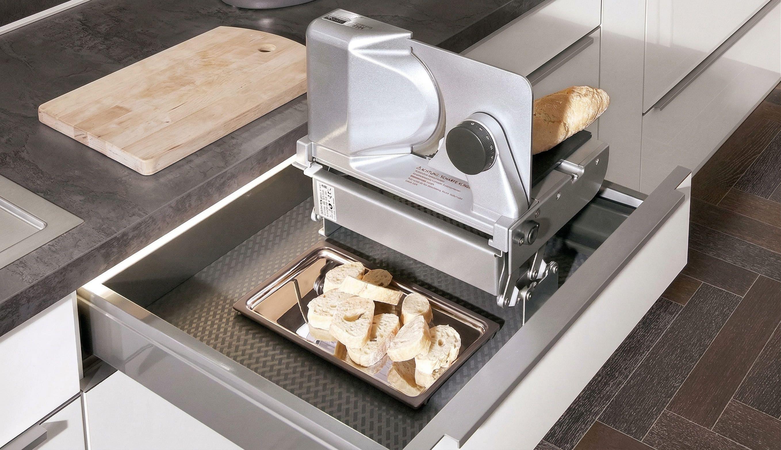 Ausziehbare Arbeitsplatte mit eingebautem Aufschnittgerät und Backblech mit geschnittenem Brot in moderner Küche.