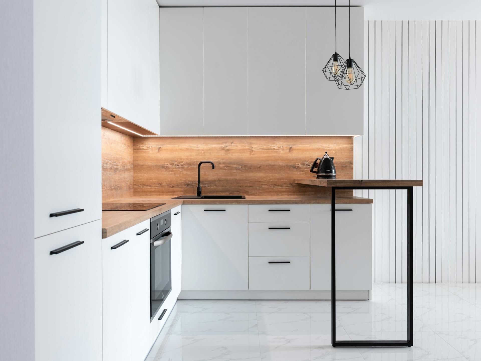 Moderne weiße Küche mit Holzrückwand, schwarzem Wasserhahn, eingebautem Ofen und zwei geometrischen Hängeleuchten über einer Holzarbeitsplatte mit schwarzem Metallgestell.